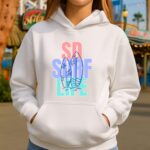 Woman wearing a white SD Surf Life hoodie with pastel surfboard design at a San Diego boardwalk